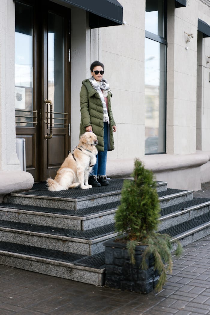 Adult woman with visual impairment stands outdoors with guide dog, wearing sunglasses and winter clothing.